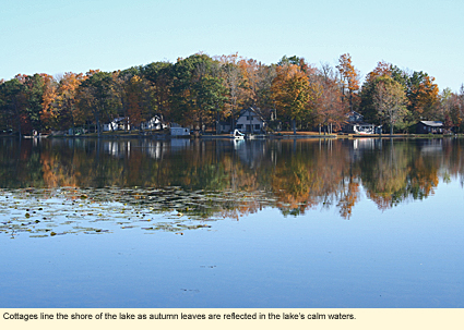Cottages line the shore of the lake as autumn leaves are reflected in the lake's calm waters.
